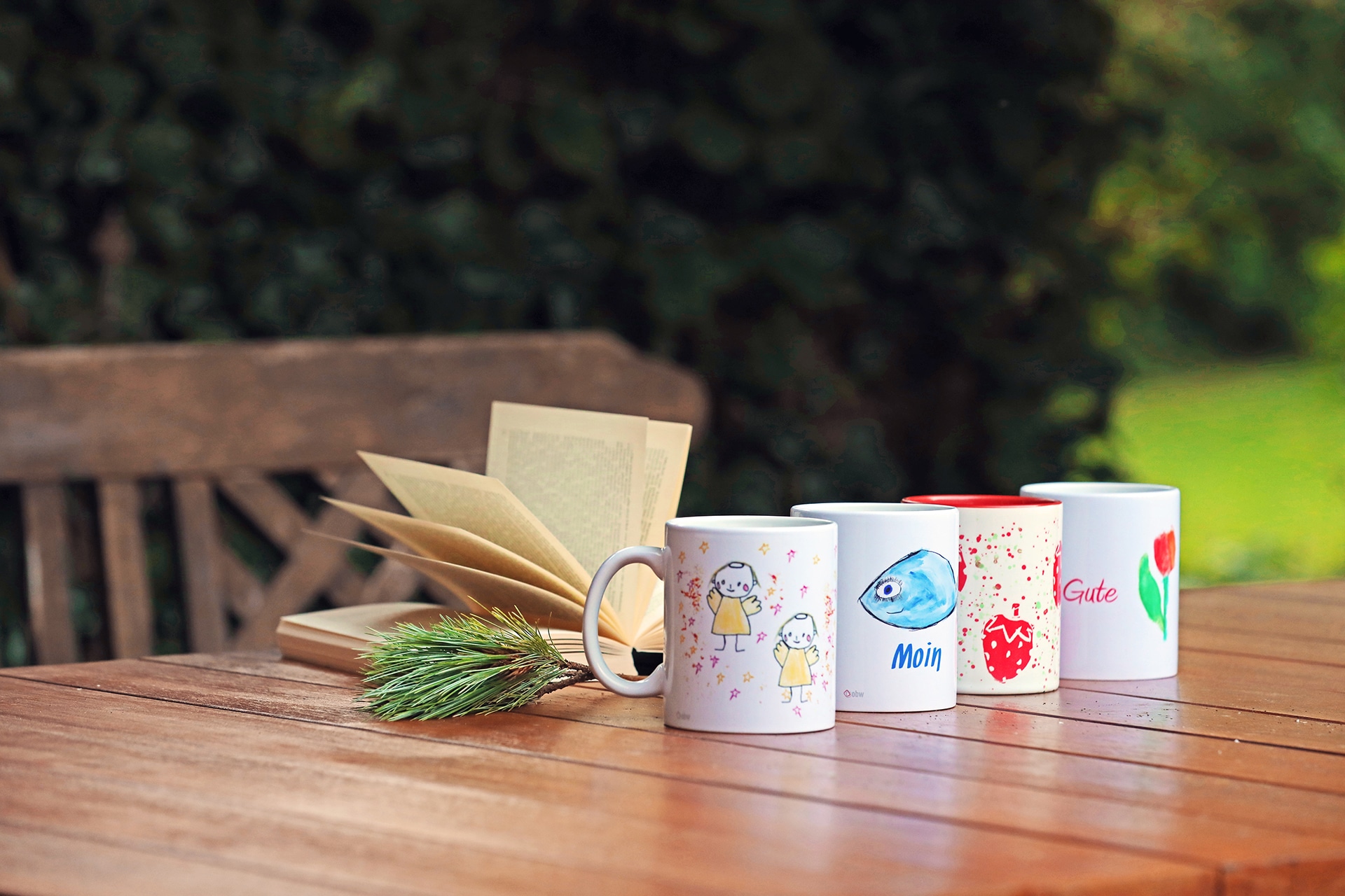Vier Tassen, bedruckt mit Engeln, Fischen, Erdbeeren und Tulpen stehen auf einem Holztisch im Garten.