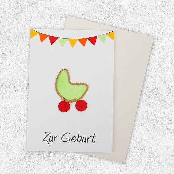 Eine Karte mit bunten Fähnchen und einer Wiege in grün mit der Aufschrift Zur Geburt mit Umschlag.
