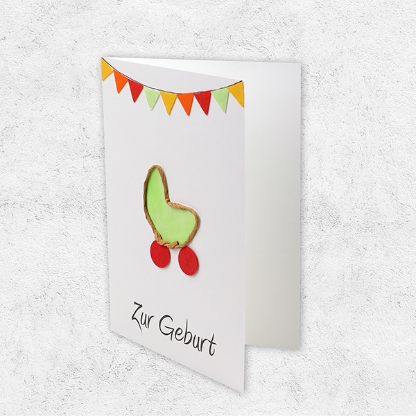Eine Karte mit bunten Fähnchen und einer Wiege in grün mit der Aufschrift Zur Geburt, seitlich.
