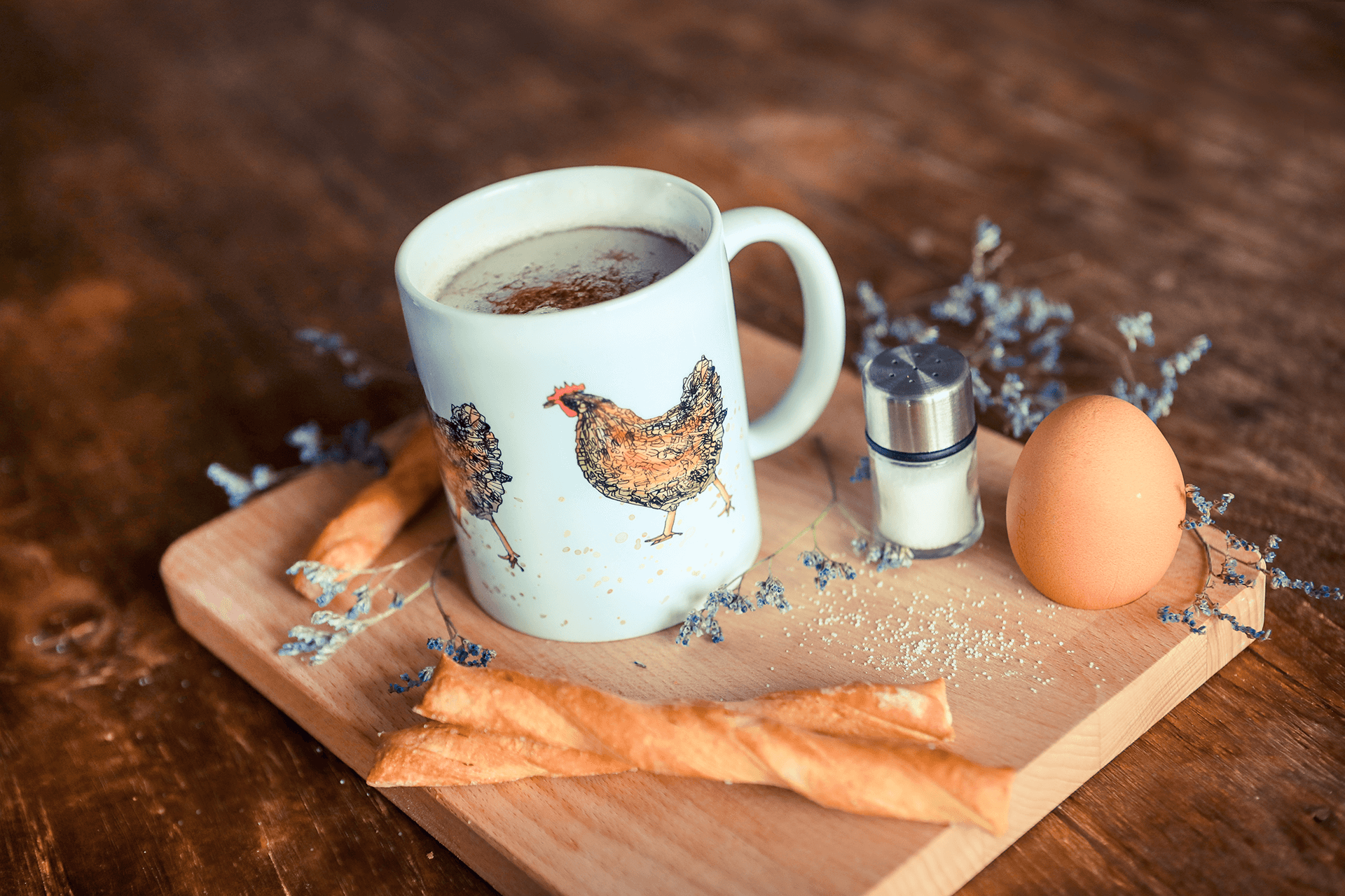 Eine Tasse, bedruckt mit dem Motiv Huhn steht auf einem Holzbrett, dekoriert mit Frühstücksei, Salzstreuer und Crackern.