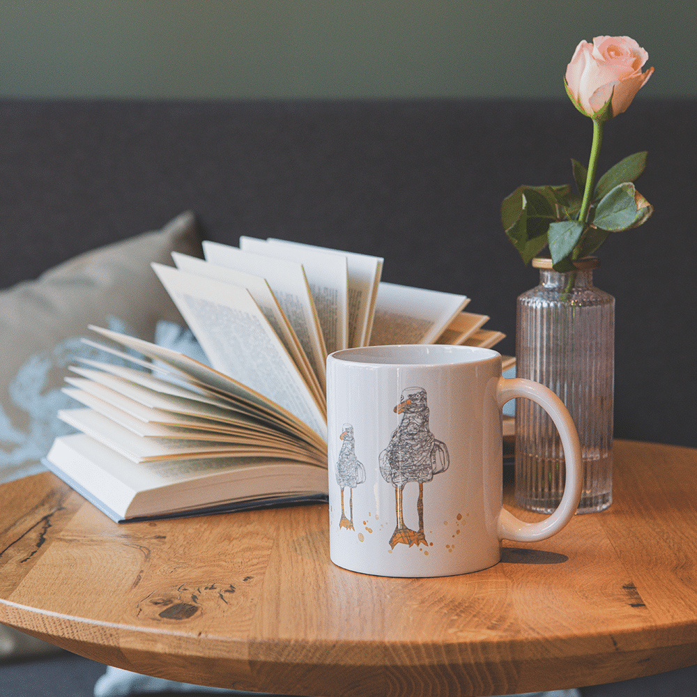 Tasse auf Holztisch mit Motiv der drei kunstvollen Möwen. Dahinter ein offenes Buch und eine Vase mit einer Rose.