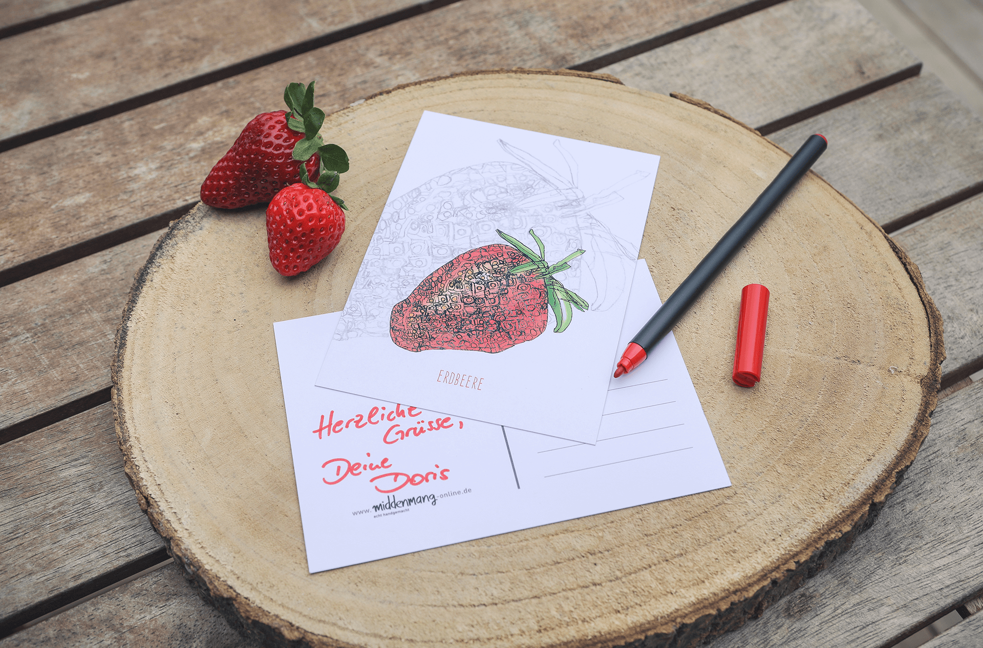 Zwei Postkarten mit dem Motiv Erdbeere auf einer Holzscheibe neben frischen Erdbeeren.