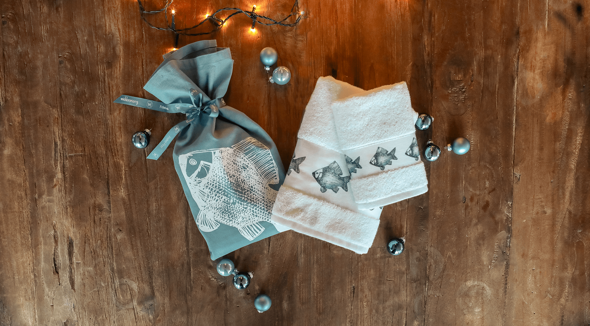 Geschirrtucher in der Farbe blau, mit dem Motiv eines Fischs, als weihnachtliches Geschenk zusammengeschnürt und weihnachtlich dekoriert, liegt neben zwei Handtücher, bedruckt mit Fischen.