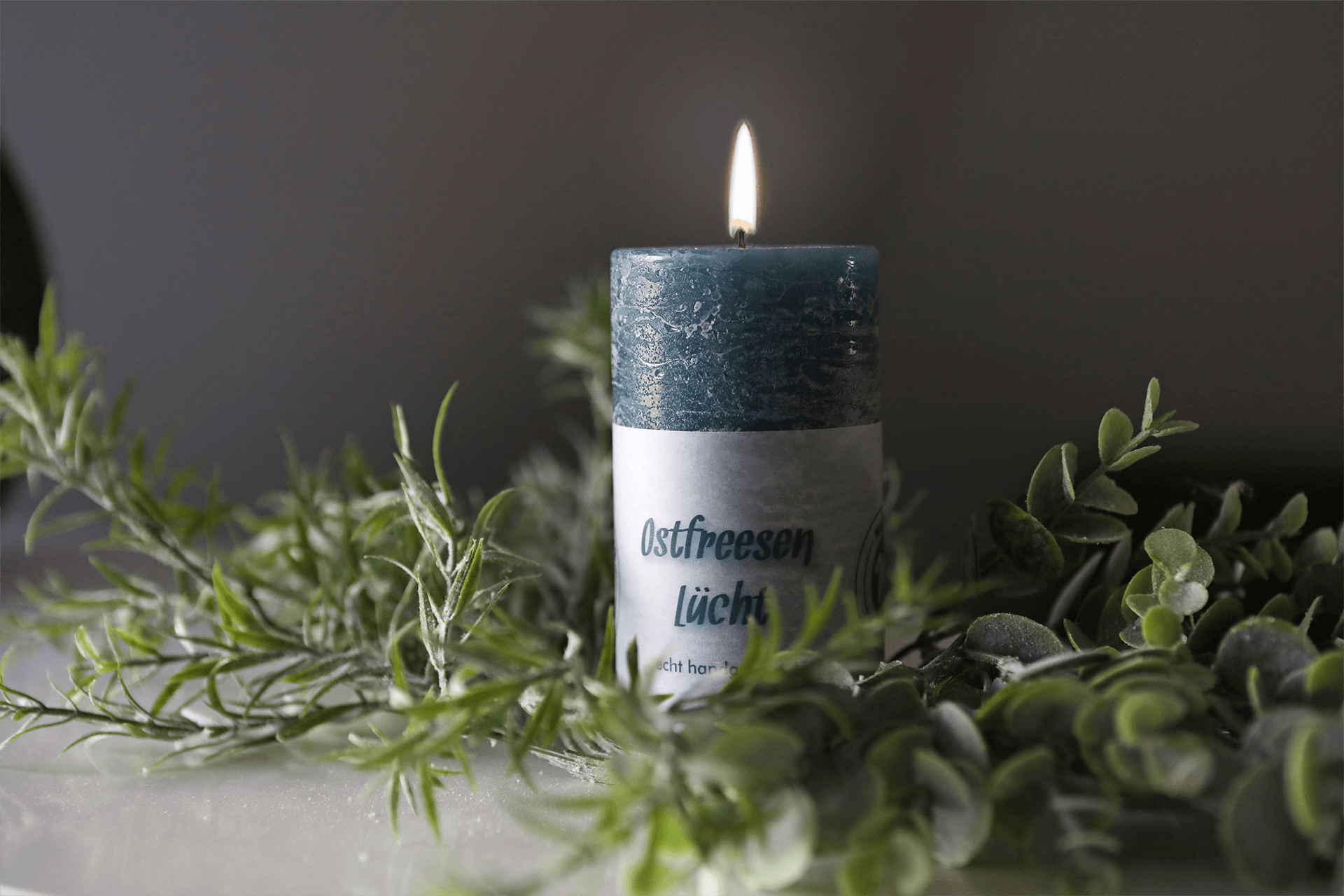 Eine runde Kerze in der Farbe dark coean mit der Aufschrift Ostfreesen Lücht echt handgemacht, dekoriert mit grünen Zweigen.