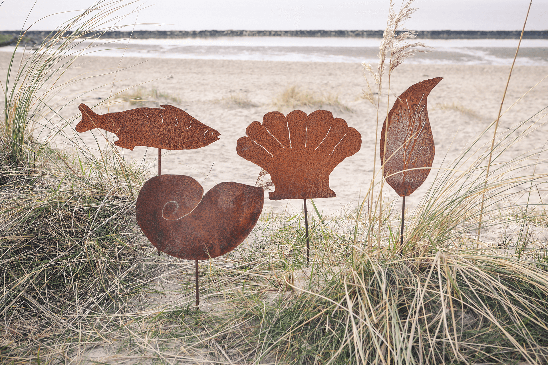 Vier Gartenstecker aus Metall in Rostoptik in den Varianten Fisch, Schnecke, Muschel und Feder am Strand.