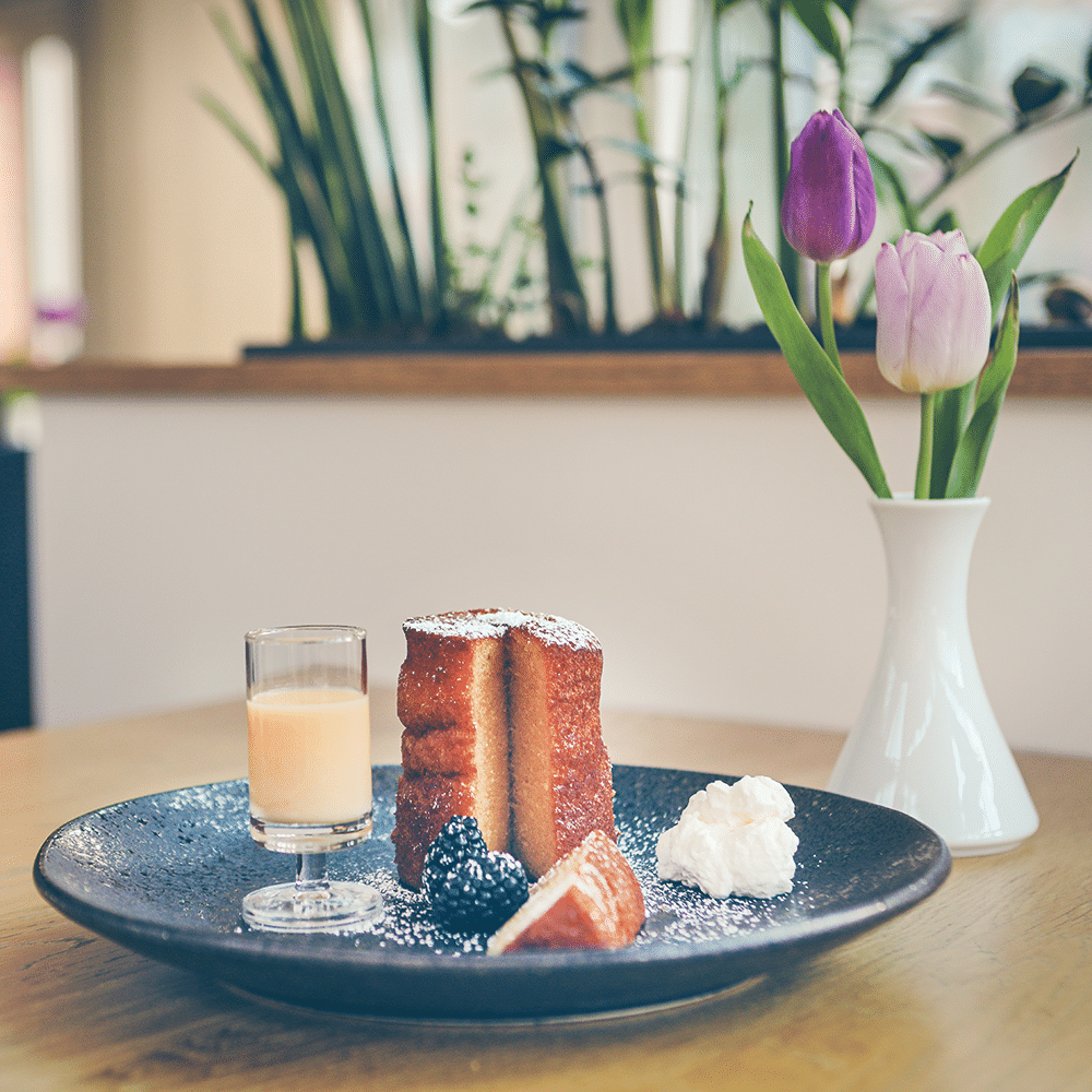 Eierlikörkuchen, angeschnitten und auf einem Teller angerichtet, mit Puderzucker, Sahne, Beeren und einem Glas Eierlikör.