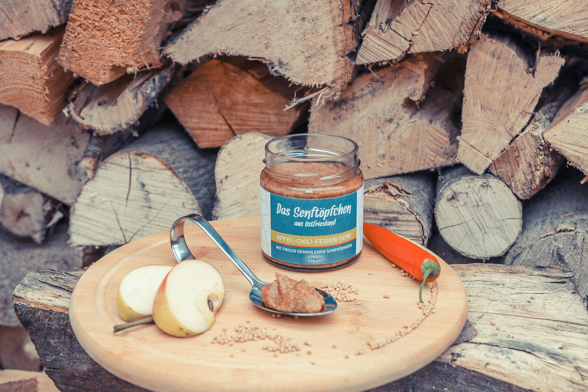 Ein geöffnetes Glas Apfel-Chili-Feigen-Senf auf einem runden Holzbrett, dekoriert mit Chili und Apfelstücken.