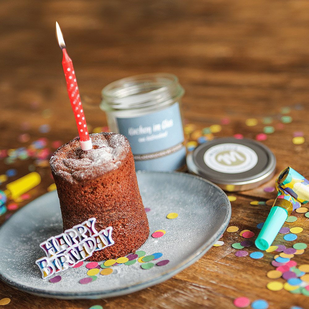 Schokoladenkuchen mit Schuss, auf einem Teller mit Kerze, Konfetti und einem Schriftzug Happy Birthday angerichtet.