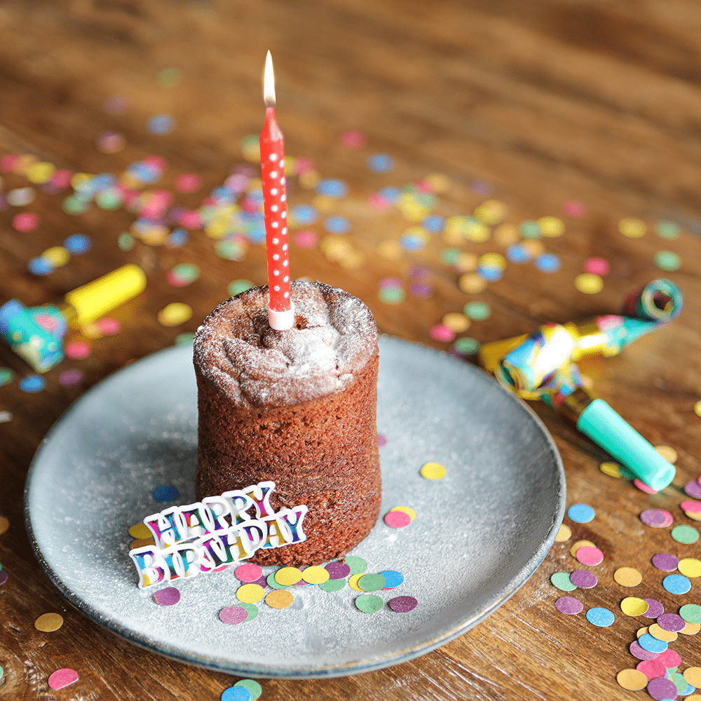 Kuchen im Glas, auf einem Teller mit einer Kerze angerichtet, Konfetti und einem Schriftzug Happy Birthday