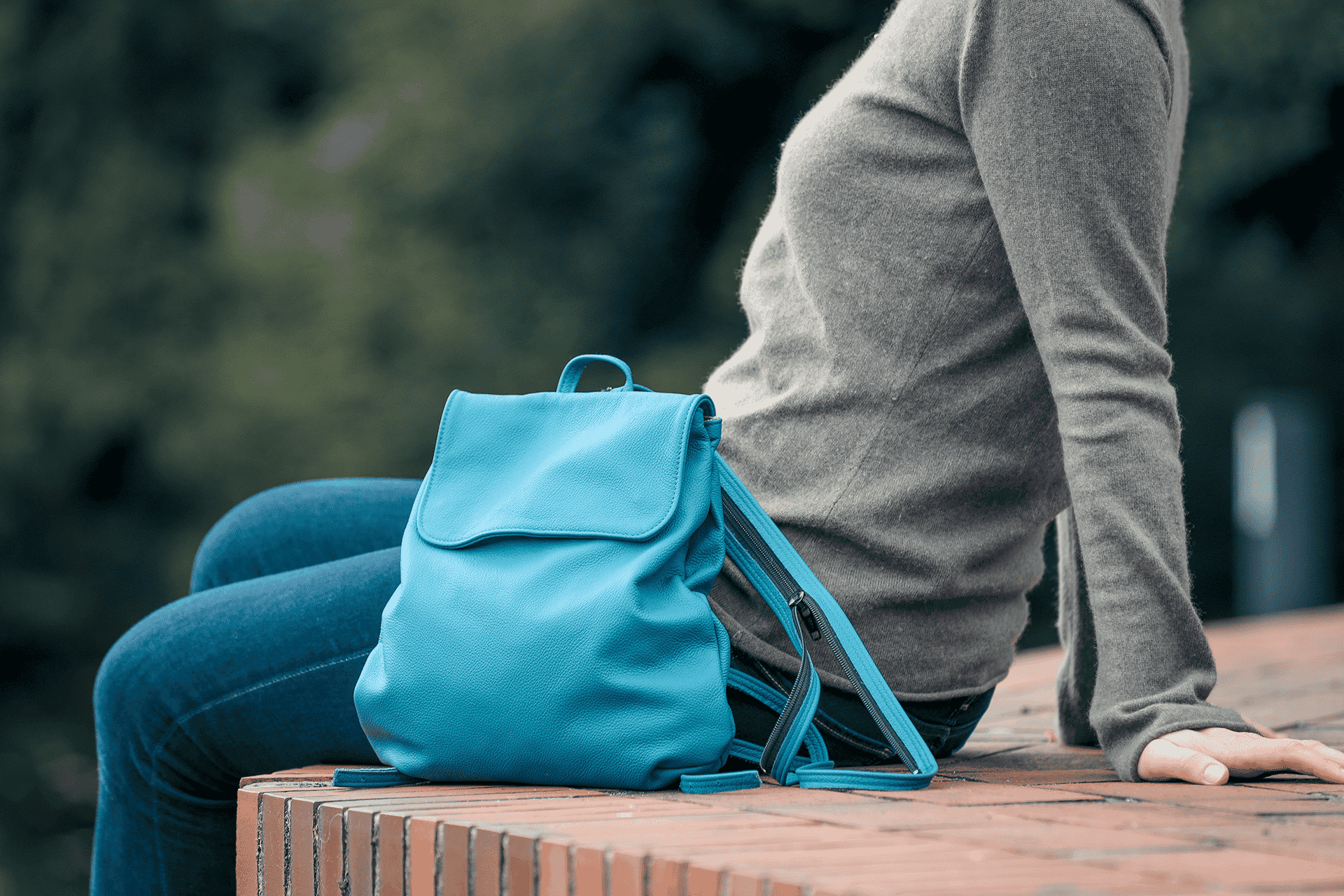 Eine Person sitzt neben einem kleinen Rucksack aus Leder in der Farbe rauchblau auf einer Mauer.