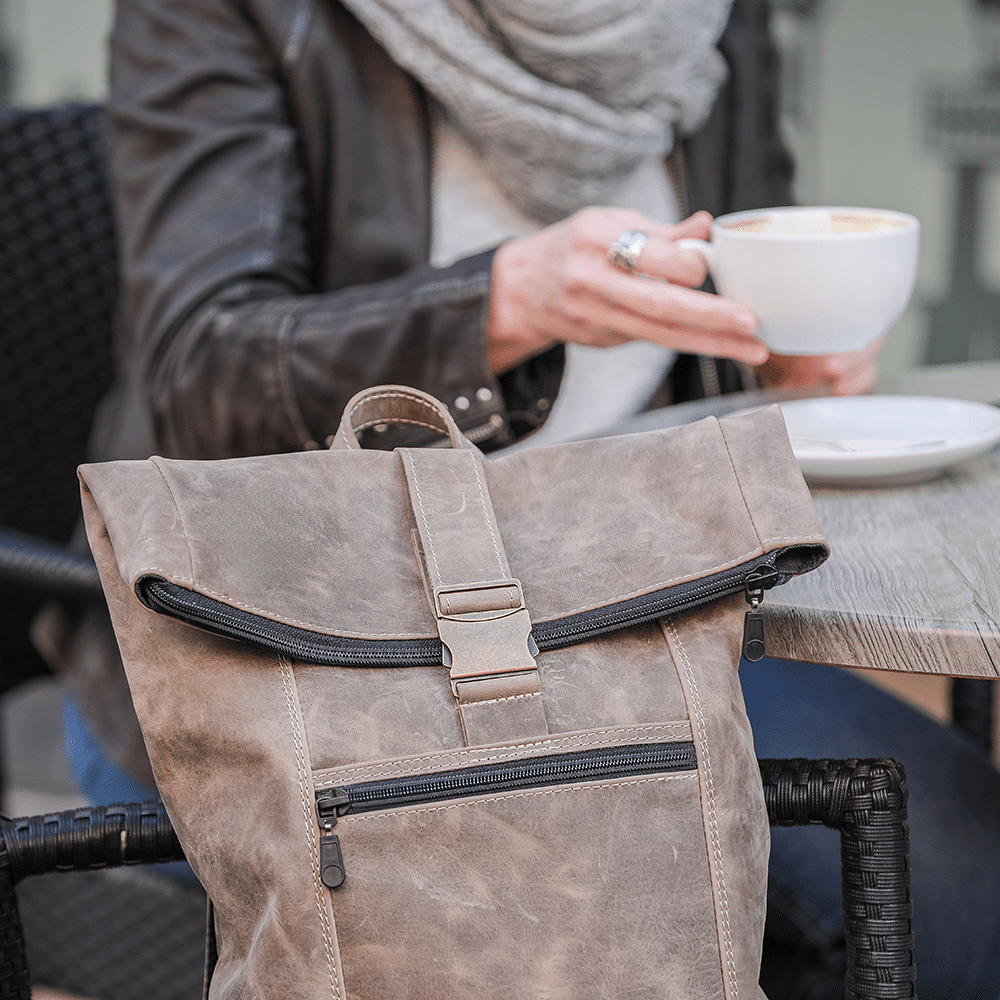 Auf einem Stuhl abgestellter Leder-Rucksack in beige und eine Person im Hintergrund, die Café trinkt.
