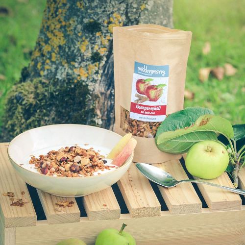 Knuspermüsli Apfel-Zimt steht im Garten auf Holzkiste neben Äpfeln und einer Schale Müsli.