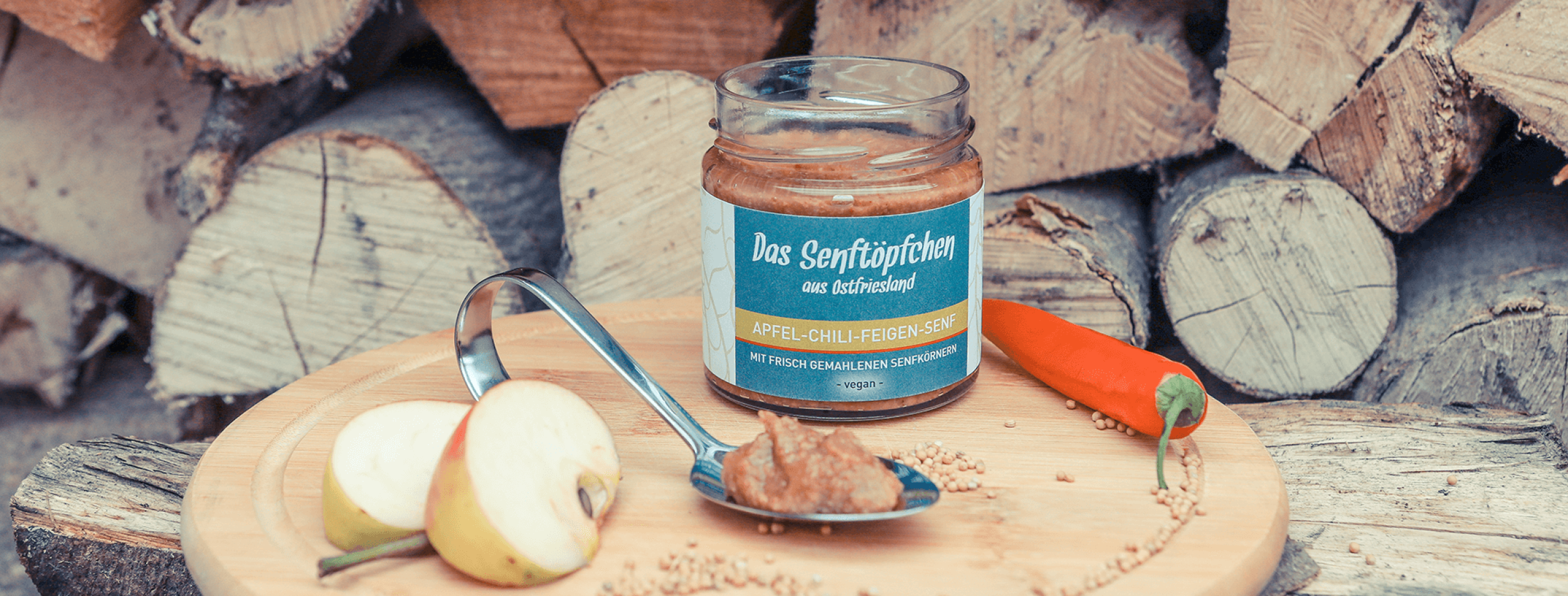 Senftöpfchen Senfglas mit orangenem Inhalt und blauem Etikett auf einem Holzbrett umgeben von einem Löffel, Apfel, Chili und Senfkörnern.