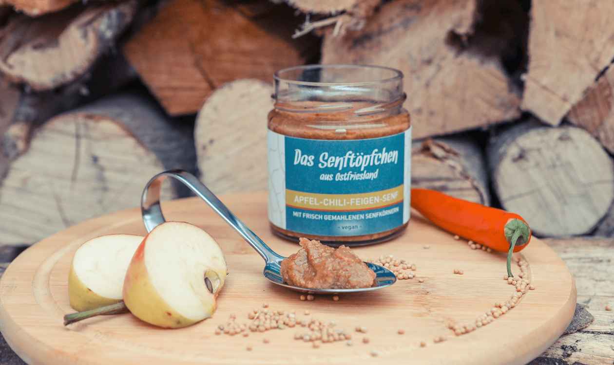 Ein geöffnetes Glas Apfel-Chili-Feigen-Senf auf einem runden Holzbrett, dekoriert mit Chili und Apfelstücken.