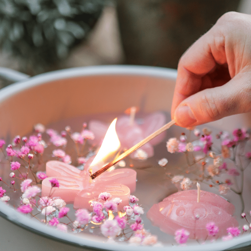 Drei Schwimmkerzen in Form von Biene, Käfer und Libelle, in der Farbe Skin rose, schwimmend in einer Schale mit Blüten. Eine Person entzündet eine Kerze.